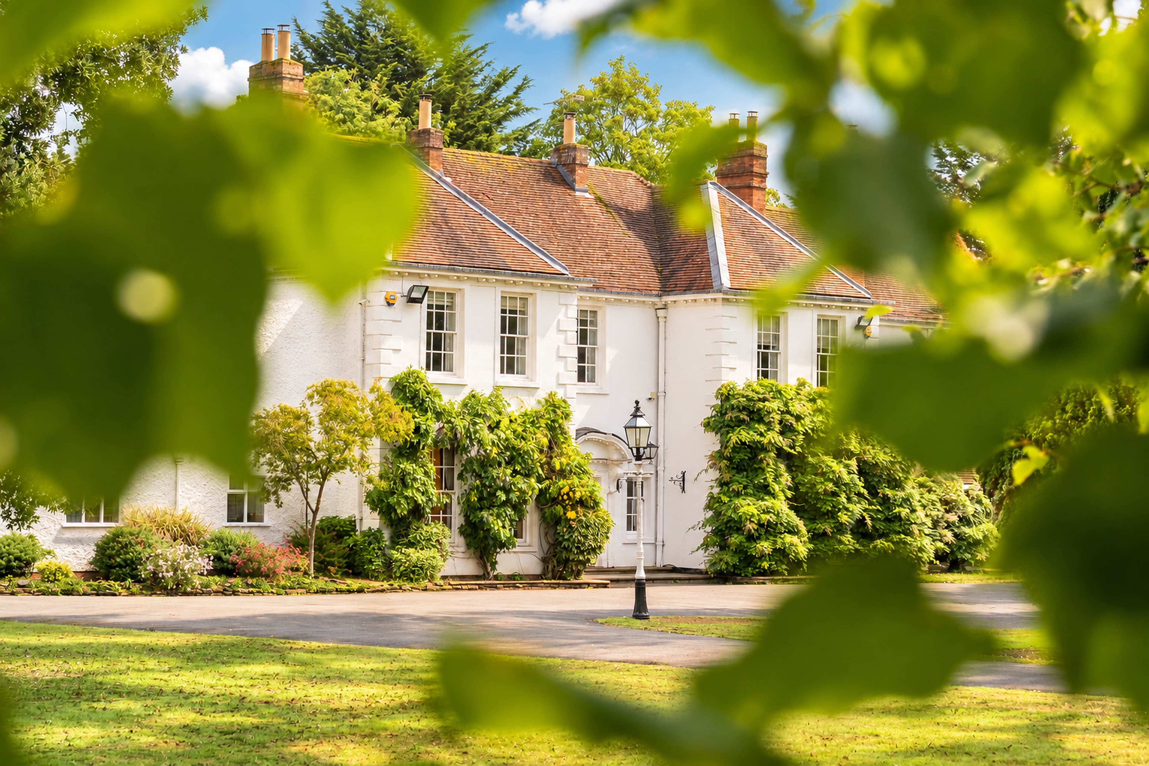 Longbridge Manor seen through soft-focus green leaves, with its front entrance and courtyard lantern visible beyond