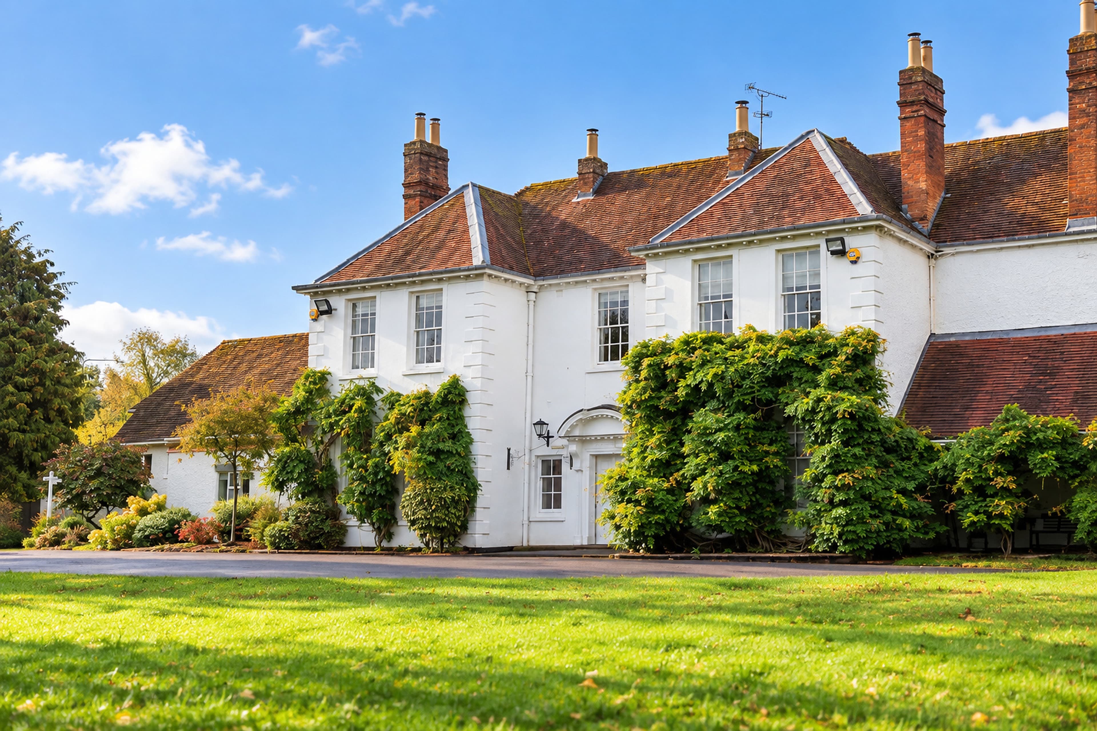 An angled view of Longbridge Manor’s white façade and climbing greenery, with the lawn in the foreground and a blue sky above