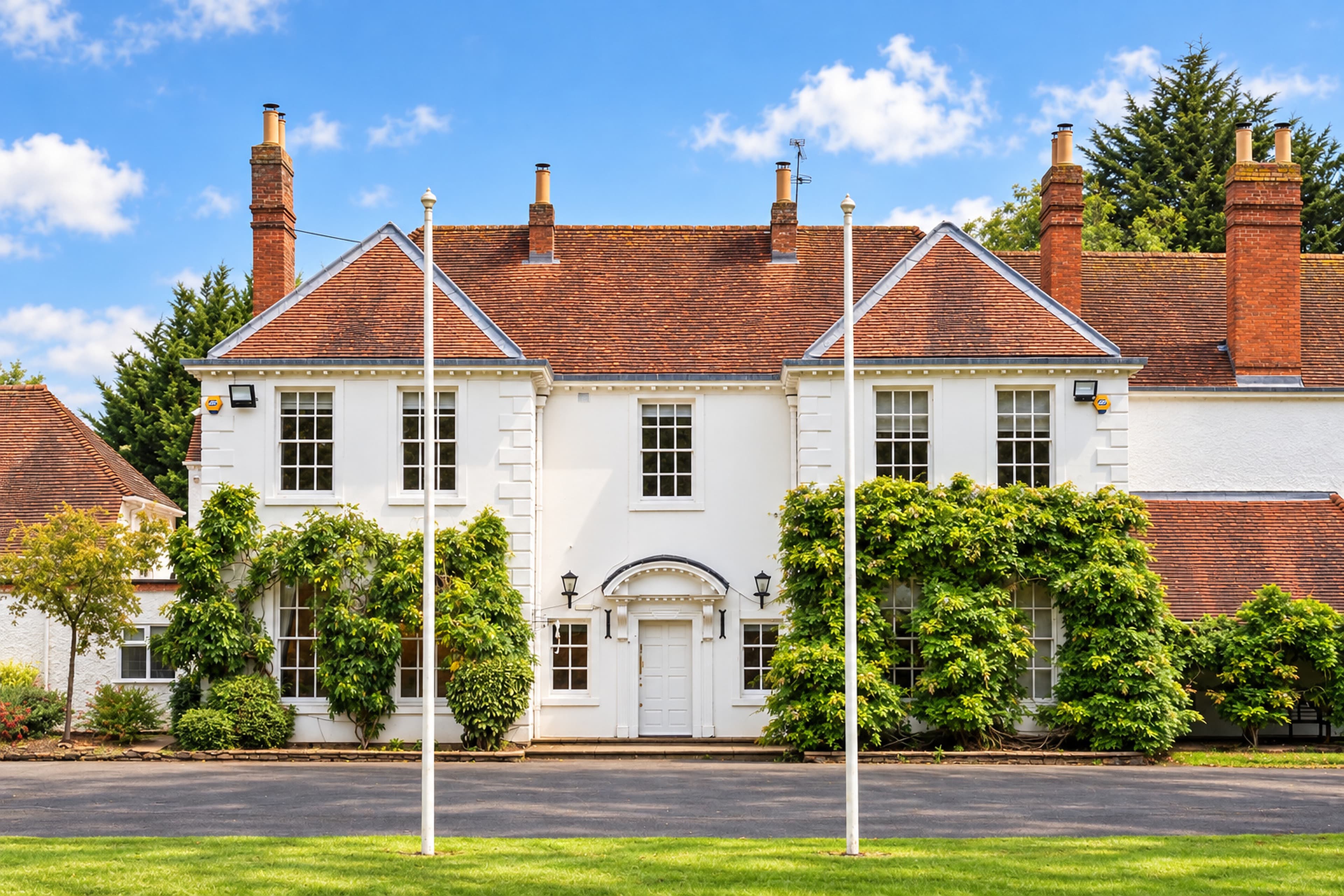 A straight-on view of Longbridge Manor’s front entrance, framed by two flagpoles, sash windows and climbing greenery