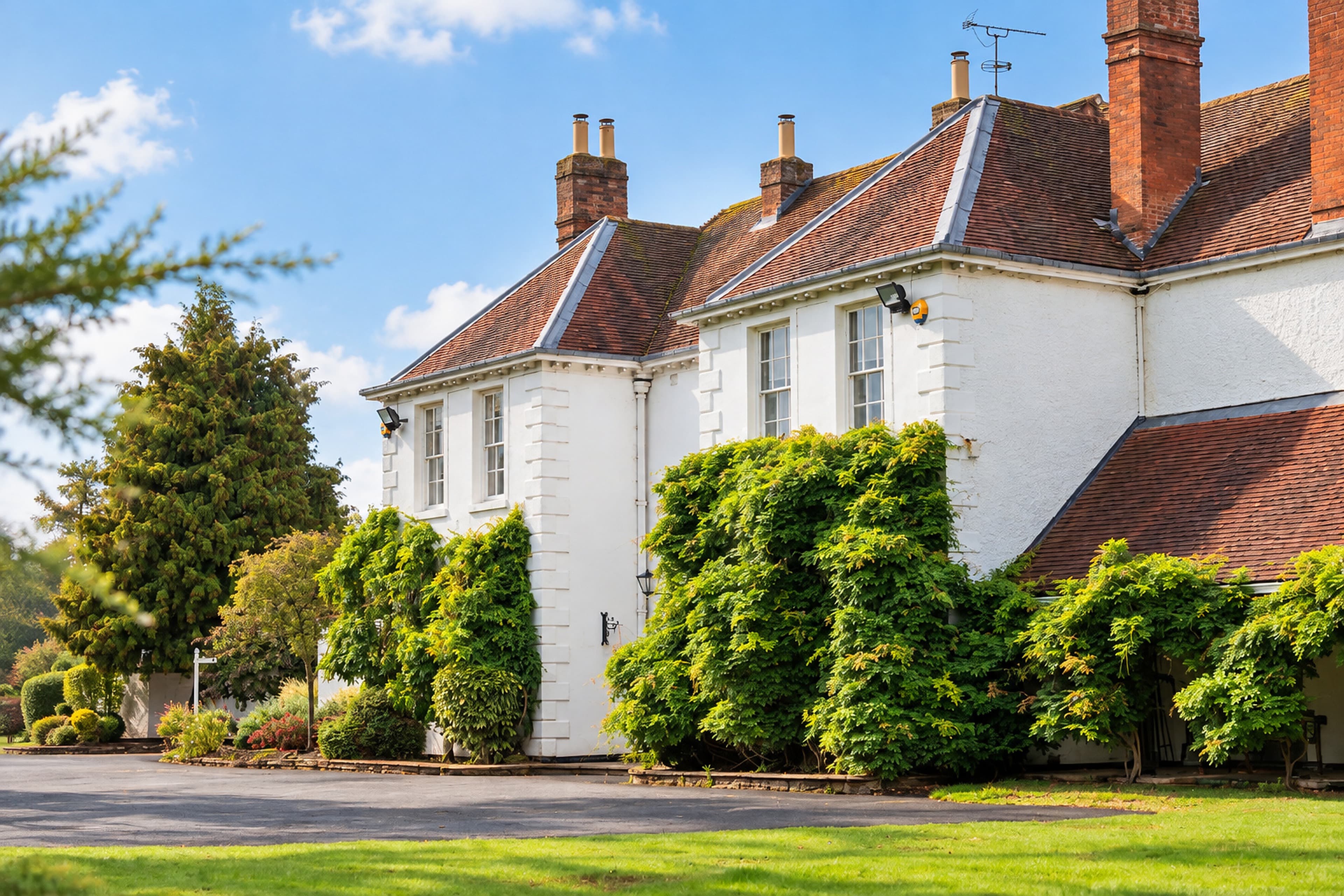 An angled view of Longbridge Manor’s red-tiled rooflines, brick chimneys and climbing greenery beside the driveway